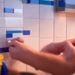 A focused close-up of a determined person’s hands carefully applying a sleek, modern peel-and-stick backsplash tile to a kitchen wall, bathed in warm cinematic lighting with rich textures and shallow depth of field highlighting the smooth adhesive surface and precision of the DIY upgrade in progress.