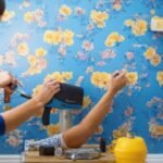 A determined homeowner’s hands carefully smoothing a stylish peel-and-stick wallpaper onto a brightly lit accent wall in a modern room, surrounded by scattered tools like a measuring tape, utility knife, and patterned wallpaper rolls, captured in cinematic lighting with rich colors and shallow depth of field to emphasize the focused DIY transformation moment