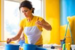 A focused shot of a determined homeowner mid-action painting a bright interior wall with a roller, surrounded by open paint cans, painter’s tape, and drop cloths, bathed in warm cinematic lighting with shallow depth of field capturing textured brush strokes and the satisfying transformation of the space, conveying empowerment and budget-friendly DIY success.