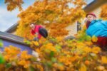 A determined homeowner wearing gloves and safety goggles climbs a sturdy ladder beside a suburban house, hands actively scooping wet leaves and debris from a clogged gutter under warm, cinematic lighting with rich colors and shallow depth of field, capturing the focused effort and moment of hands-on DIY home maintenance against a softly blurred background