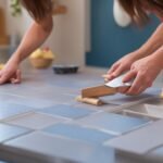 A focused close-up of determined hands carefully applying sleek peel-and-stick tiles to a kitchen backsplash in warm, cinematic lighting, with scattered DIY tools like a utility knife and smoothing tool subtly visible, capturing the satisfying moment of a modern, budget-friendly home transformation in progress.