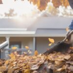 A determined homeowner wearing work gloves carefully cleaning clogged gutters on a sturdy ladder outdoors, surrounded by autumn leaves and DIY tools like a garden trowel and bucket, captured with cinematic lighting, dramatic low angle, intense focus on hands clearing debris, rich natural colors, and shallow depth of field to evoke hands-on effort and empowerment in routine home maintenance.